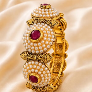 May include: A gold hinged bangle bracelet featuring three circular clusters of white pearls. Each cluster is centered with a faceted deep red gemstone. The gold metalwork displays intricate scroll patterns between the pearl-encrusted sections.