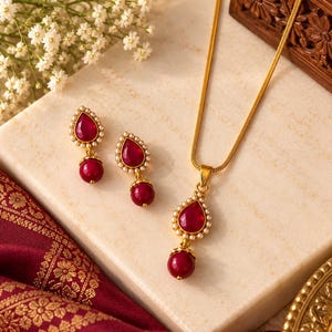 May include: A gold-toned jewelry set consisting of a necklace and a pair of drop earrings. Each piece features a teardrop-shaped deep red stone surrounded by small white pearls, with a round red bead dangling from the bottom of each pendant.