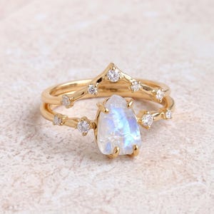 May include: A gold ring set consisting of a chevron-shaped band and a second band featuring a pear-shaped moonstone centre stone. Both bands are accented with small, round, clear stones set along the gold metal shanks, creating a layered look.