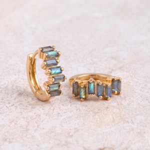 Puede incluir: Un par de pendientes de aro dorados con una hilera de piedras preciosas rectangulares de labradorita iridiscente, dispuestas verticalmente en la parte frontal de cada aro. Las piedras muestran destellos de luz azul y verde sobre el metal dorado.