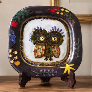 May include: A square ceramic plate with a dark purple border and a white center featuring a stylized owl illustration. The border is decorated with small, colorful painted motifs including red, yellow, and blue shapes, resting on a dark wooden stand.