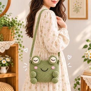 May include: A green crochet crossbody bag shaped like a frog with closed eyes, pink cheeks, and a black smile. The bag features a long matching crochet strap and two small rounded feet at the bottom, with white heart illustrations drawn on the sides.