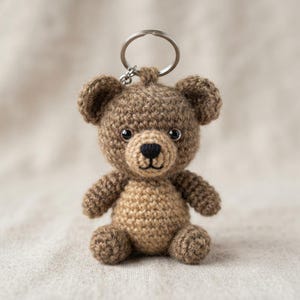 Può includere: Un piccolo portachiavi a forma di orsetto all'uncinetto con testa e orecchie marrone scuro e un muso beige più chiaro. L'orsetto ha occhi in plastica nera e naso e bocca ricamati in nero. Un anello in metallo color argento è fissato alla testa.