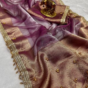 May include: A sheer mauve fabric drape featuring a wide, ornate gold metallic border with intricate floral patterns and dangling gold beads. The fabric is accented with small, scattered gold embellishments and a gold cow figurine ornament.