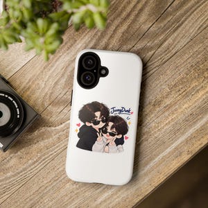 May include: A white smartphone case featuring a cartoon illustration of two people with dark hair wearing sunglasses and hoodies. The text JoongDunk is printed in blue above the figures, accompanied by small red hearts and yellow star icons.