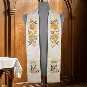 May include: A cream-colored liturgical stole featuring gold embroidered roses with blue leaves on the upper section and a gold crown above a blue and gold letter M monogram on the lower section, displayed on a gray mannequin torso.