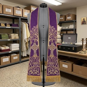 May include: A purple liturgical stole featuring gold embroidered Celtic cross designs, intricate tree of life motifs, and knotwork patterns. The garment is finished with gold fringe at the bottom edges and is displayed on a gray dress form mannequin.