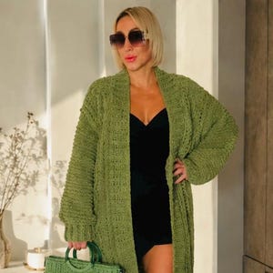 Pode incluir: Um cardigã longo de malha trançada em verde-oliva com frente aberta e mangas compridas, combinado com um vestido curto preto e botas de couro castanho, acompanhado por uma mala de mão entrançada verde-oliva com pegas redondas e alça preta fina.