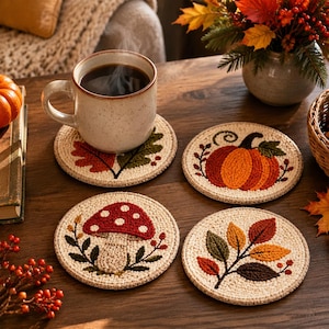 Könnte beinhalten: Ein Set aus vier runden, cremefarbenen Untersetzern mit strukturierten, gestickten Herbstmotiven: ein rot-oranger Kürbis, ein rot-weißer Pilz, ein mehrfarbiger Blattzweig und ein einzelnes Eichenblatt mit detaillierten Stichen.