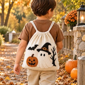 May include: A cream-colored drawstring backpack featuring a textured, embroidered Halloween design. The artwork includes a smiling orange jack-o'-lantern, a white ghost wearing a black witch hat, a small black bat, and a bare black tree branch.