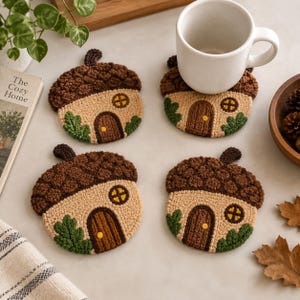 May include: Four round, textured coasters shaped like acorns, each featuring a tan house design with a brown door, a yellow window, and green leaves. The top of each coaster has a dark brown, bumpy cap texture. One coaster supports a white mug.