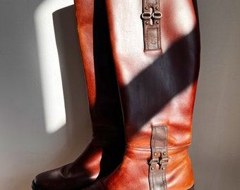 May include: A pair of knee-high brown leather boots. The boots have a classic design with a buckle detail and a flat sole. The leather has a rich, warm tone, and the boots appear to be in good condition.