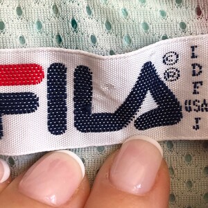 May include: A white label with the FILA logo in navy blue and red. The label includes size information and the text "MADE IN ITALY". The background is a light green perforated fabric.