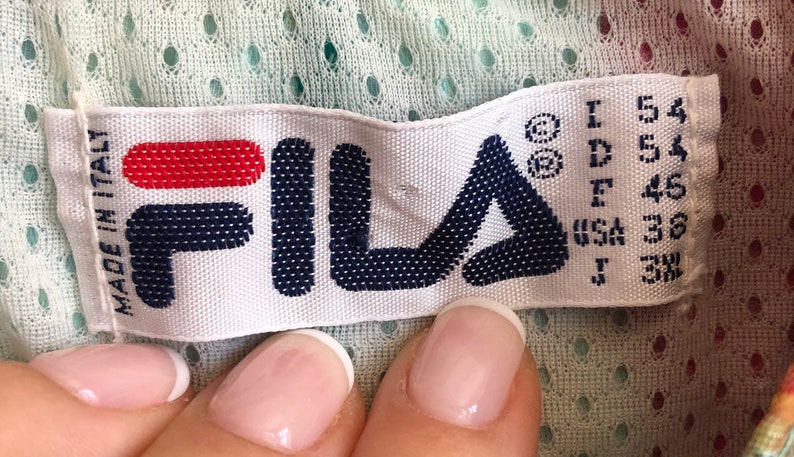 May include: A white label with the FILA logo in navy blue and red. The label includes size information and the text "MADE IN ITALY". The background is a light green perforated fabric.