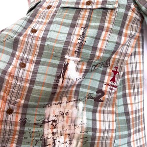 May include: A button-down plaid shirt in shades of green, brown, and orange. The shirt features two chest pockets and brown buttons. The fabric has a distressed look with faded text and graphics. The shirt has the text "ARAFANO 196" and the number "2".