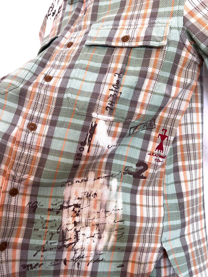 May include: A button-down plaid shirt in shades of green, brown, and orange. The shirt features two chest pockets and brown buttons. The fabric has a distressed look with faded text and graphics. The shirt has the text "ARAFANO 196" and the number "2".
