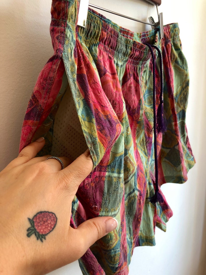 May include: Colorful patterned shorts with a drawstring waist and a small pocket. The shorts feature a vibrant mix of pink, green, and purple hues. The shorts are hanging on a wooden hanger.