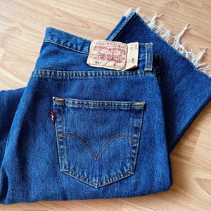 Pode incluir: Jeans de ganga azul escuro com um design clássico de cinco bolsos. Os jeans apresentam uma etiqueta vermelha Levi's no bolso traseiro e um patch de couro. A bainha crua adiciona um toque moderno ao estilo de inspiração vintage.