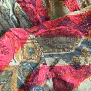 May include: Close-up of a colorful fabric with a patchwork design. The fabric features a mix of pink, red, blue, and gold geometric patterns. Visible stitching adds detail to the design. The overall theme is textile art.