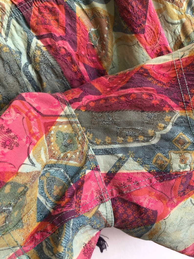 May include: Close-up of a colorful fabric with a patchwork design. The fabric features a mix of pink, red, blue, and gold geometric patterns. Visible stitching adds detail to the design. The overall theme is textile art.