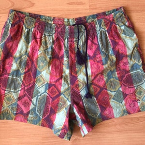 May include: Colorful patterned shorts with an elastic waistband and a drawstring with tassels. The shorts feature a repeating geometric design in shades of pink, red, blue, and gold.