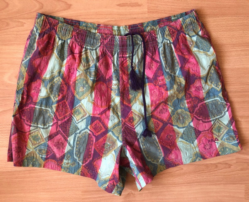 May include: Colorful patterned shorts with an elastic waistband and a drawstring with tassels. The shorts feature a repeating geometric design in shades of pink, red, blue, and gold.