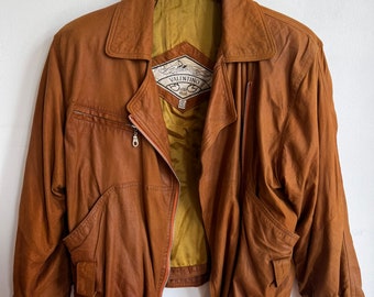 May include: A brown leather jacket with a zip closure, side pockets, and a collared neckline. The jacket is hanging on a wooden hanger against a white background. The interior lining is a golden color.