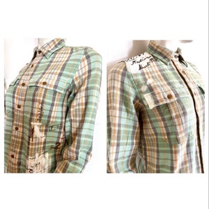 May include: A long-sleeved plaid shirt in shades of green, brown, and white. The shirt features two chest pockets and button closures. Handwritten text and numbers are visible on the front and shoulder of the shirt.