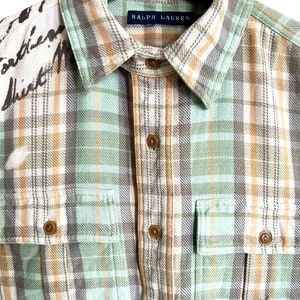 May include: A button-down plaid shirt in shades of green, tan, and gray. The shirt features two chest pockets and a collar with the "Ralph Lauren" label. The left shoulder has handwritten text.