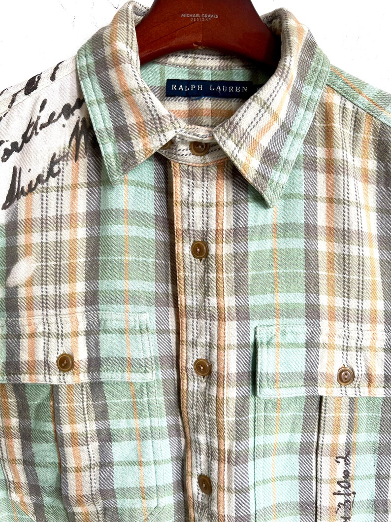 May include: A button-down plaid shirt in shades of green, tan, and gray. The shirt features two chest pockets and a collar with the "Ralph Lauren" label. The left shoulder has handwritten text.