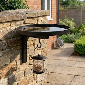 May include: A black metal wall-mounted bird bath featuring a circular basin filled with water. A matching black metal bird feeder with a wire mesh cylinder containing birdseed hangs from a hook attached to the underside of the bird bath bracket.