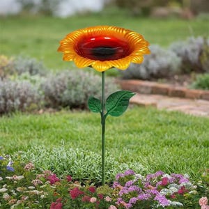 May include: A sunflower-shaped bird bath featuring a vibrant yellow and orange petal rim surrounding a deep red basin. The basin is supported by a thin green metal stem with two green leaves attached, standing upright in a garden setting.