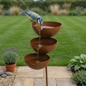 Puede incluir: Una fuente de agua de tres niveles con tres cuencos de metal apilados de color óxido. El agua fluye desde el cuenco superior a través de los cuencos central e inferior. Un pequeño pájaro azul, amarillo y blanco bebe del borde superior.