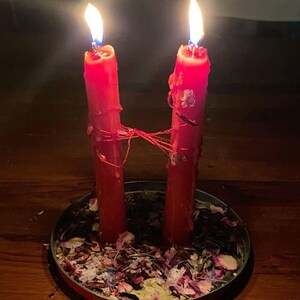 May include: Two tall red pillar candles with lit flames, connected by a thin red string. The candles stand upright in a shallow circular metal tray filled with a mixture of dried flower petals, herbs, and small botanical fragments.