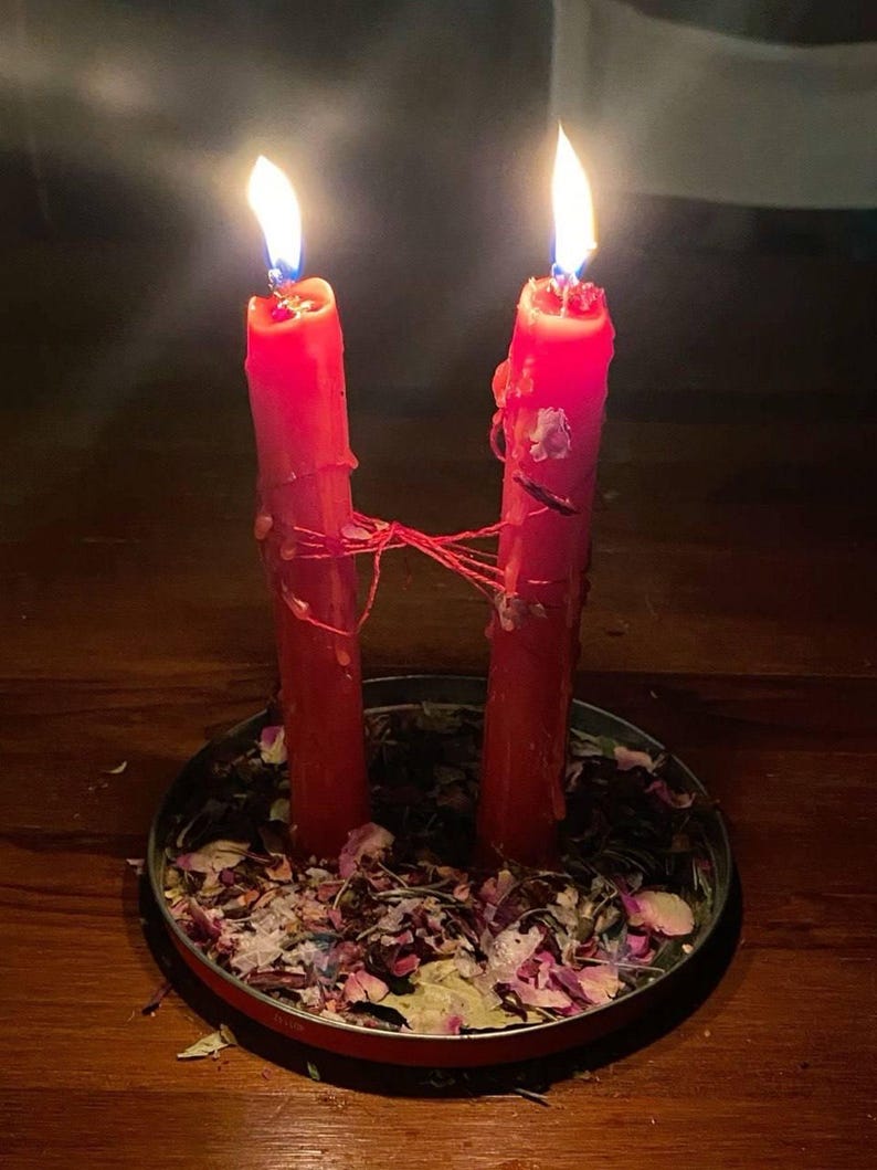 May include: Two tall red pillar candles with lit flames, connected by a thin red string. The candles stand upright in a shallow circular metal tray filled with a mixture of dried flower petals, herbs, and small botanical fragments.