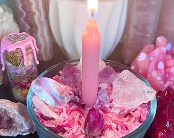 May include: A lit pink taper candle centered in a clear glass bowl filled with pink and white rice, dried rosebuds, and various raw crystals including clear quartz and amethyst. The candle flame is bright, casting light on the surrounding stones.