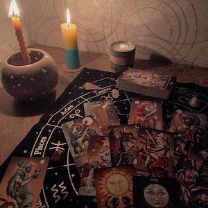 May include: A black fabric cloth featuring gold zodiac signs and constellations, spread with a deck of tarot cards. Two lit candles, one in a round ceramic holder and one bicolor yellow and blue, sit nearby alongside a small grey cylindrical container.