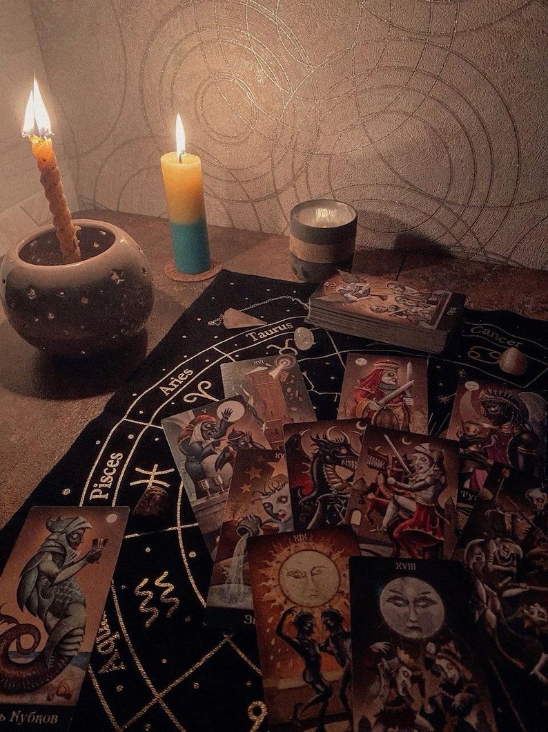 May include: A black fabric cloth featuring gold zodiac signs and constellations, spread with a deck of tarot cards. Two lit candles, one in a round ceramic holder and one bicolor yellow and blue, sit nearby alongside a small grey cylindrical container.