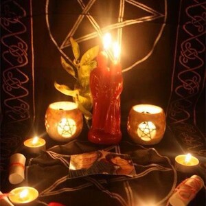 May include: A ritual setup featuring a black cloth with a white pentagram, a red dual-figure candle, two stone candle holders with pentagram cutouts, several tea light candles, and rolled paper scrolls tied with red ribbon, all illuminated by warm light.
