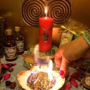 May include: A bright red pillar candle with a silver spiral pendant sits behind a burning seashell filled with dried herbs. Two glass apothecary bottles with labels reading Romero and Limon are visible alongside scattered red petals and small stones.