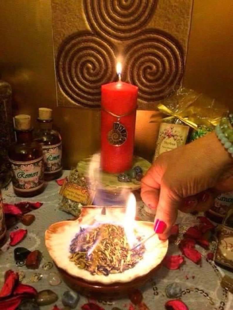 May include: A bright red pillar candle with a silver spiral pendant sits behind a burning seashell filled with dried herbs. Two glass apothecary bottles with labels reading Romero and Limon are visible alongside scattered red petals and small stones.