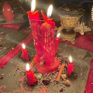 May include: Two red candles bound together with red string, both lit with visible flames. They are surrounded by four smaller lit red candles, cinnamon sticks, and scattered dried herbs on a dark grey stone slab surface.
