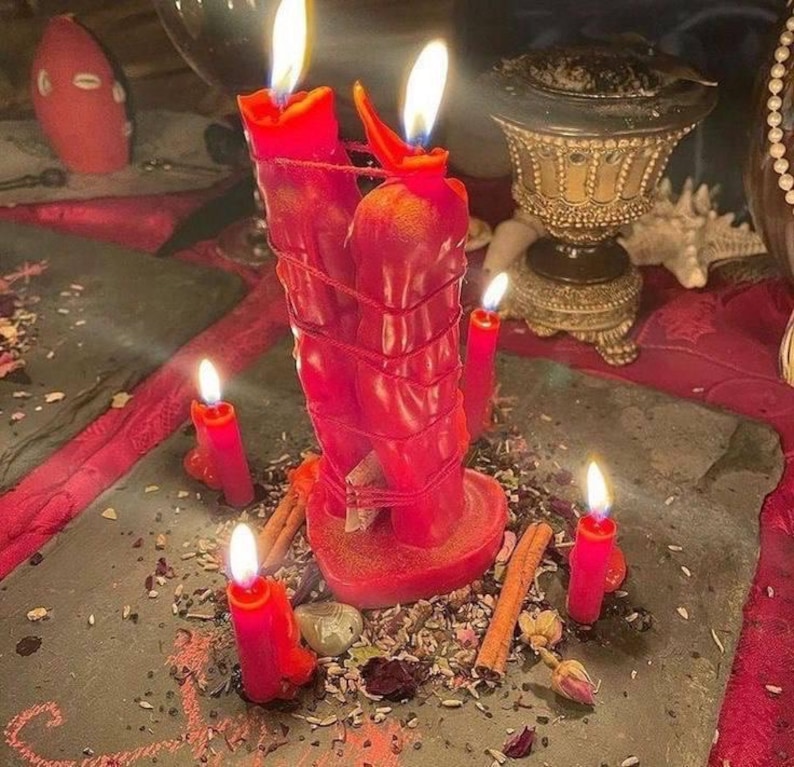 May include: Two red candles bound together with red string, both lit with visible flames. They are surrounded by four smaller lit red candles, cinnamon sticks, and scattered dried herbs on a dark grey stone slab surface.