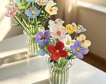Könnte beinhalten: Zwei klare Glasvasen mit vertikaler Riffelung, die jeweils einen Strauß kleiner, mehrfarbiger Glasblumen enthalten. Die Blumen haben Blütenblätter in den Farben Rot, Blau, Gelb, Violett, Rosa und Weiß sowie grüne Glasstiele und gelbe Mitten.