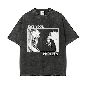 Könnte beinhalten: Ein anthrazitfarbenes T-Shirt in Acid-Wash-Optik mit einem weißen, rechteckigen Grafikdruck. Das Motiv zeigt das Profil einer Person mit offenem Mund und einer Hand, die Nahrung hält, sowie den Text EAT YOUR PROTEIN in abgenutzter Schrift.