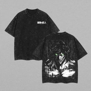 Può includere: Una t-shirt nera con effetto lavaggio acido, caratterizzata da un piccolo logo con testo giapponese bianco sul petto. Il retro della maglietta mostra una grande e dettagliata illustrazione grafica in bianco e nero di un personaggio con occhi verdi luminosi e denti affilati.