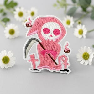 Puede incluir: Un parche bordado que muestra una figura rosa con capucha, cara de calavera blanca y cuencas oculares rosas. La figura sostiene una guadaña gris y se encuentra entre dos pequeñas cruces rosas, con un pequeño remolino de humo rosa a la derecha.