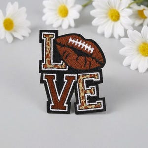 Puede incluir: Un parche bordado con la palabra LOVE en letras de bloque. La L y la E tienen un patrón de leopardo marrón, mientras que la O está reemplazada por un gráfico de labios con forma de balón de fútbol marrón con cordones blancos.