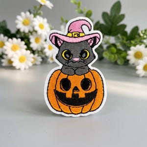 Puede incluir: Un parche bordado que muestra un gato gris con grandes ojos amarillos que lleva un sombrero de bruja rosa, sentado sobre una calabaza naranja con una cara tallada en negro. El diseño tiene un borde blanco y está hecho con hilo detallado.