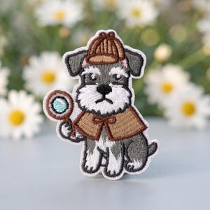 May include: An embroidered patch featuring a gray and white schnauzer dog dressed as a detective. The dog wears a brown deerstalker hat and a matching brown cape, while holding a small magnifying glass with a light blue lens in its paw.